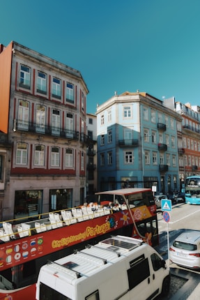 A vibrant city street scene features colorful old buildings with ornate facades, a red open-top tourist bus advertising city sightseeing in Portugal, and various vehicles parked and driving along the cobblestone street. The sky is clear and bright blue.