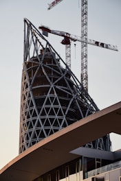 A high-rise building under construction with a unique geometric metal framework on its top section. Several cranes are visible, towering over the structure and indicating ongoing construction work. The base of the building features modern architecture with glass windows.