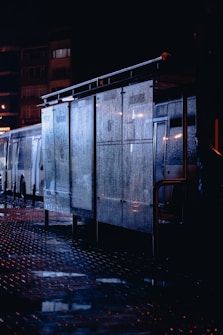 Rain-soaked street scene featuring a dimly lit bus stop with glass panes covered in droplets, reflecting the surrounding city lights. The pavement is wet, creating reflective surfaces under the darkened sky. A parked bus is partially visible in the background, with buildings faintly outlined.