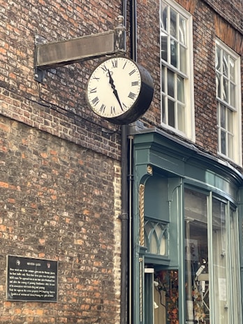 A vintage clock with Roman numerals is mounted on a brick wall above a shop entrance. The clock shows the time as approximately ten minutes to ten. The shop has a classic design with teal-colored woodwork and large glass windows. A historical plaque is attached to the brick wall beneath the clock.