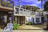 A two-story beach house with a white and wooden exterior. The upper floor features a balcony with a striped umbrella and outdoor seating. There are potted plants and greenery around the ground level, which includes large windows and a wooden deck area.
