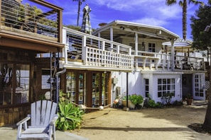 A two-story beach house with a white and wooden exterior. The upper floor features a balcony with a striped umbrella and outdoor seating. There are potted plants and greenery around the ground level, which includes large windows and a wooden deck area.