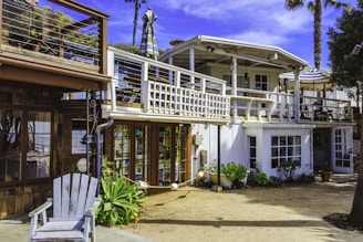 A two-story beach house with a white and wooden exterior. The upper floor features a balcony with a striped umbrella and outdoor seating. There are potted plants and greenery around the ground level, which includes large windows and a wooden deck area.