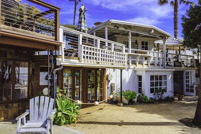 A two-story beach house with a white and wooden exterior. The upper floor features a balcony with a striped umbrella and outdoor seating. There are potted plants and greenery around the ground level, which includes large windows and a wooden deck area.