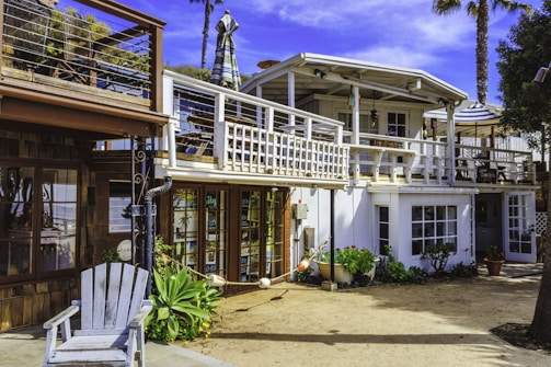 A two-story beach house with a white and wooden exterior. The upper floor features a balcony with a striped umbrella and outdoor seating. There are potted plants and greenery around the ground level, which includes large windows and a wooden deck area.