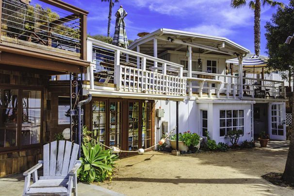 A two-story beach house with a white and wooden exterior. The upper floor features a balcony with a striped umbrella and outdoor seating. There are potted plants and greenery around the ground level, which includes large windows and a wooden deck area.