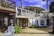 A two-story beach house with a white and wooden exterior. The upper floor features a balcony with a striped umbrella and outdoor seating. There are potted plants and greenery around the ground level, which includes large windows and a wooden deck area.