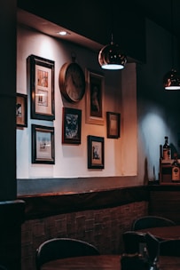 Several framed pictures and a large round wall clock decorate a dimly lit interior wall, creating a cozy ambiance. Copper hanging lights provide soft illumination, highlighting the textures of the wall and the wooden elements of the room.