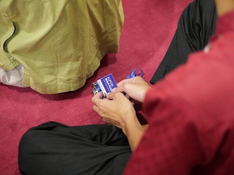 Two individuals are sitting on a red carpet, with one holding a blue coupon that has text on it. The individual’s clothing includes a red garment, and another person is wearing green fabric.