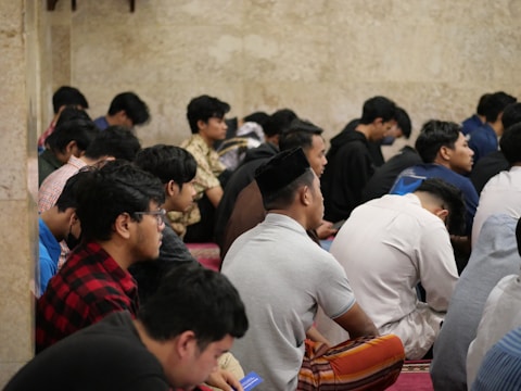 A group of people seated closely together in a contemplative or prayerful posture. The environment appears to be indoors with a neutral-colored stone wall in the background.