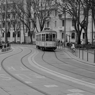 An old-fashioned tram is traveling along curved tracks on a street lined with leafless trees and historic buildings. Several people stand nearby, and bicycles are parked against a railing.