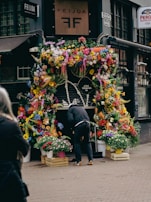 A storefront is lavishly decorated with a vibrant array of flowers in various colors including yellow, pink, orange, and purple. There are numerous potted plants placed on wooden crates and white containers. A person is bending down near the flowers in the center, possibly arranging them. The surroundings include a dark building facade with signage displaying the name 'Feijoa' and another sign indicating a direction. The setting appears to be on a cobblestone street with another person partially visible on the left.