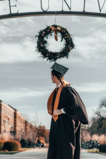 A person in graduation attire, wearing a cap and gown with an orange sash, stands under a decorative archway adorned with a wreath. The backdrop includes a clear sky and the architecture of a university campus.