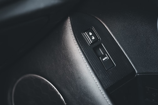 A close-up of a car's interior showing a door panel with a window control switch and door lock buttons. The panel surface is textured with a grainy pattern, and the buttons feature icons for lock and unlock, as well as an auto button for windows.