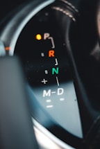 Close-up view of a car's gear shift indicator with visible symbols for park, reverse, neutral, and drive modes. The indicator lights are illuminated, and there are markings for manual shifting options.