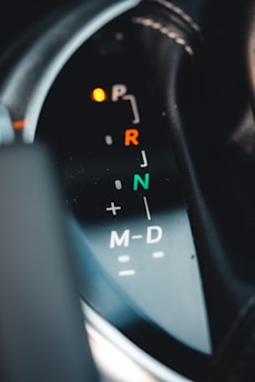 Close-up view of a car's gear shift indicator with visible symbols for park, reverse, neutral, and drive modes. The indicator lights are illuminated, and there are markings for manual shifting options.