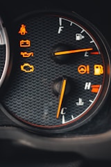 A close-up view of a car dashboard featuring multiple warning lights and gauges. The fuel gauge indicates a full tank while the temperature gauge points towards the cooler side. Various warning icons are illuminated in orange, including seatbelt, battery, oil, and engine alerts, set against a textured background.