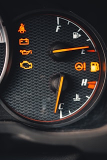 A close-up view of a car dashboard featuring multiple warning lights and gauges. The fuel gauge indicates a full tank while the temperature gauge points towards the cooler side. Various warning icons are illuminated in orange, including seatbelt, battery, oil, and engine alerts, set against a textured background.
