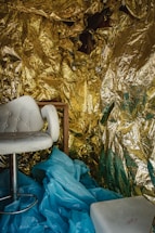 A cozy interior space is draped with gold foil, creating a shimmering, reflective background. A white cushioned chair with a chrome base sits prominently in the foreground, surrounded by a crumpled layer of light blue material on the floor. The setup exudes an eclectic mix of textures and colors.