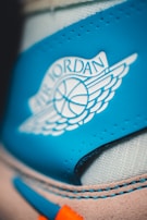 Close-up of a blue and white shoe with a visible 'Air Jordan' logo featuring a basketball and wings. The stitching and suede texture around the logo are sharply detailed.