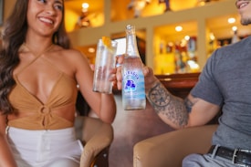 Two individuals are seated closely in a warmly lit setting, possibly a bar or cafe. One person is holding a tall glass with a slice of orange, possibly containing a drink, while the other is holding a bottle labeled 'Nicaragua.' The atmosphere is relaxed and friendly, with a focus on socializing.