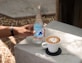 A hand wearing a black wristband holds a bottle of mineral water labeled 'Mineragua' on a concrete surface. Next to the bottle, there is a white cup of coffee with latte art placed on a black coaster. Sunlight casts shadows on the concrete table, and some dry leaves are scattered on the ground.