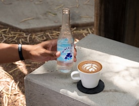 A hand wearing a black wristband holds a bottle of mineral water labeled 'Mineragua' on a concrete surface. Next to the bottle, there is a white cup of coffee with latte art placed on a black coaster. Sunlight casts shadows on the concrete table, and some dry leaves are scattered on the ground.