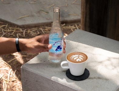 A hand wearing a black wristband holds a bottle of mineral water labeled 'Mineragua' on a concrete surface. Next to the bottle, there is a white cup of coffee with latte art placed on a black coaster. Sunlight casts shadows on the concrete table, and some dry leaves are scattered on the ground.