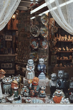 A shop window display features an array of intricately designed cat figurines. The figures, crafted in whimsical outfits and various expressions, stand on shelves. The display is framed by sheer white curtains, allowing a glimpse into the interior, where additional crafts and decorative plates are visible.
