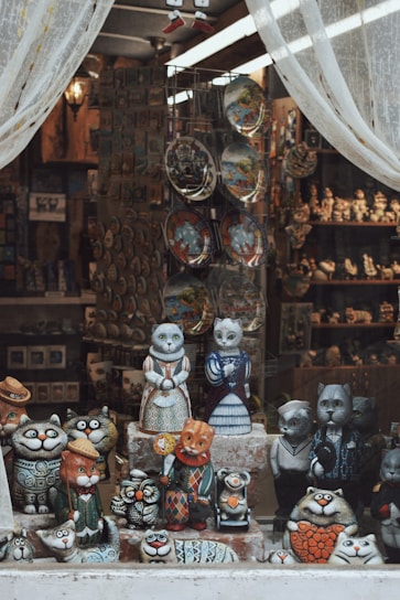A shop window display features an array of intricately designed cat figurines. The figures, crafted in whimsical outfits and various expressions, stand on shelves. The display is framed by sheer white curtains, allowing a glimpse into the interior, where additional crafts and decorative plates are visible.