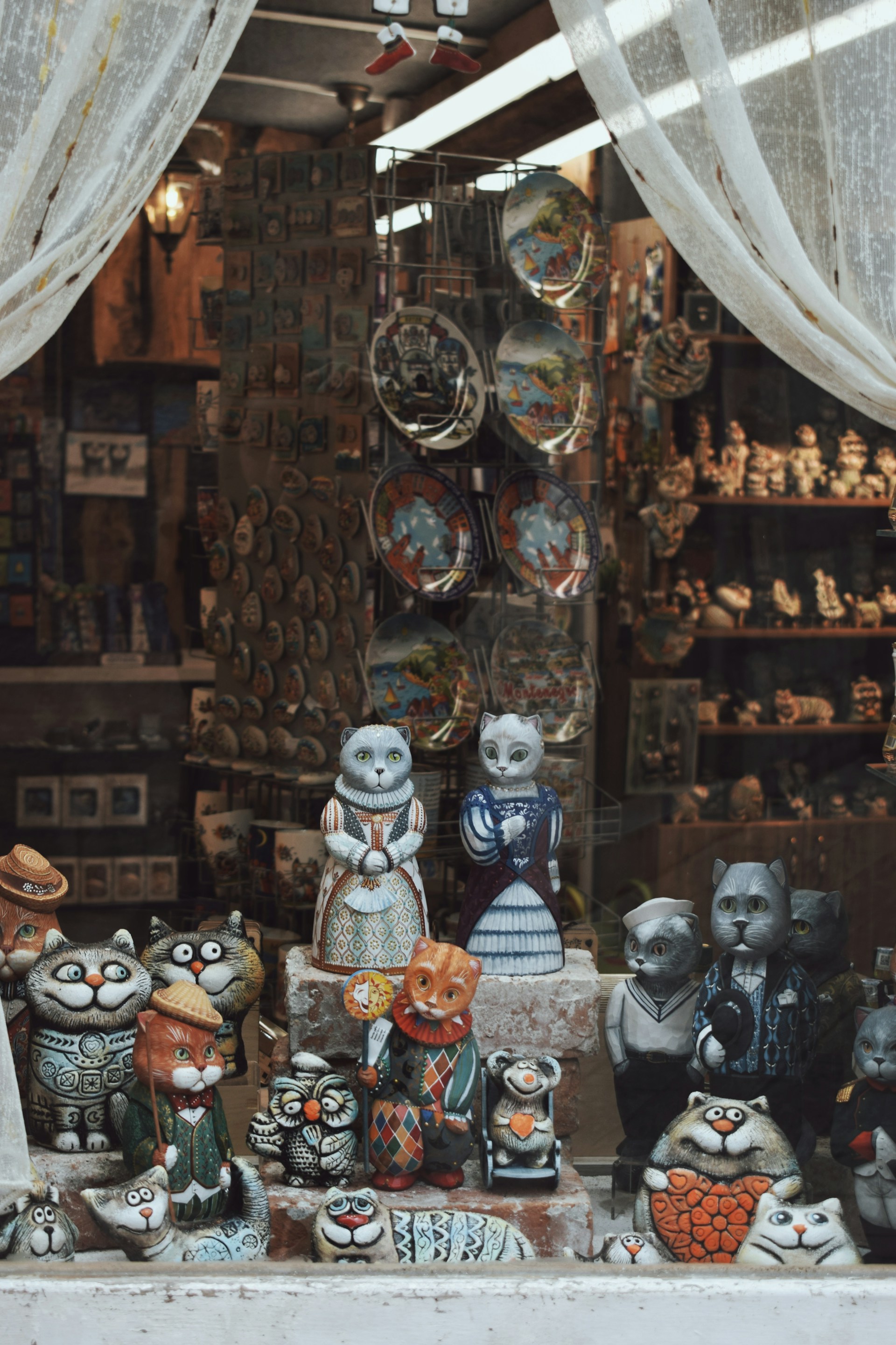 A shop window display features an array of intricately designed cat figurines. The figures, crafted in whimsical outfits and various expressions, stand on shelves. The display is framed by sheer white curtains, allowing a glimpse into the interior, where additional crafts and decorative plates are visible.
