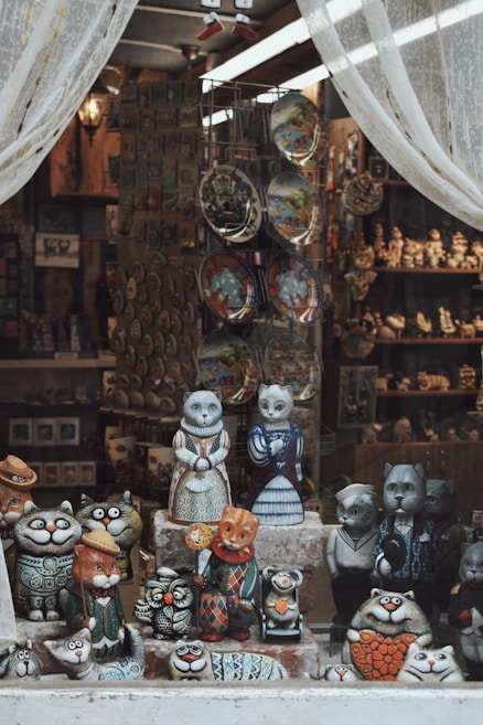 A shop window display features an array of intricately designed cat figurines. The figures, crafted in whimsical outfits and various expressions, stand on shelves. The display is framed by sheer white curtains, allowing a glimpse into the interior, where additional crafts and decorative plates are visible.