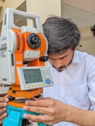 A person is intently working with a modern surveying instrument, adjusting its components. The equipment has an orange and white body, with a digital display and various controls, suggesting precision and technical usage.
