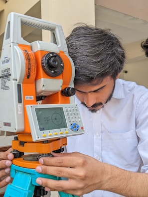 A person is intently working with a modern surveying instrument, adjusting its components. The equipment has an orange and white body, with a digital display and various controls, suggesting precision and technical usage.