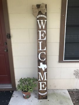 A tall wooden sign propped against the wall of a house, featuring large white letters spelling 'WELCOME' with a silhouette of Texas replacing the letter 'O'. A decorative bow is attached at the top, and a small potted plant is placed at the base of the sign.