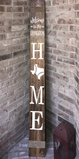A vertical wooden sign leaning against a brick corner reads 'Welcome to the Livingston Home' with the word 'HOME' vertically arranged. The letter 'O' is replaced by an outline of the state of Texas featuring a baseball stitch pattern. A decorative lantern sits on the floor beside the sign.