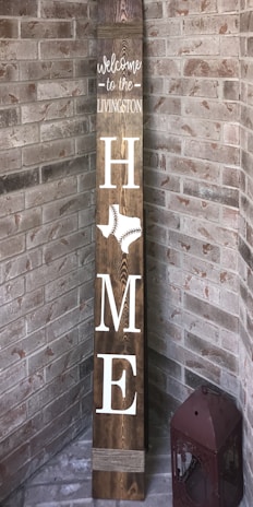 A vertical wooden sign leaning against a brick corner reads 'Welcome to the Livingston Home' with the word 'HOME' vertically arranged. The letter 'O' is replaced by an outline of the state of Texas featuring a baseball stitch pattern. A decorative lantern sits on the floor beside the sign.