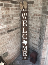 A vertical wooden welcome sign is leaning against a brick wall in a corner. The sign has the word 'WELCOME' written in white letters with the shape of Texas replacing the letter 'O'. At the top of the sign, there is a decorative beige bow. At the bottom of the sign, there are four white fleur-de-lis symbols. Next to the sign on the ground is a red, weathered lantern.
