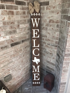 A vertical wooden welcome sign is leaning against a brick wall in a corner. The sign has the word 'WELCOME' written in white letters with the shape of Texas replacing the letter 'O'. At the top of the sign, there is a decorative beige bow. At the bottom of the sign, there are four white fleur-de-lis symbols. Next to the sign on the ground is a red, weathered lantern.