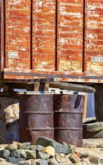 A rusty, weathered container elevated on steel drums with chipped paint and surrounded by rocks. The surface of the container indicates significant age and exposure to the elements.