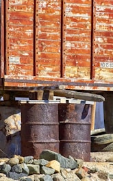 A rusty, weathered container elevated on steel drums with chipped paint and surrounded by rocks. The surface of the container indicates significant age and exposure to the elements.