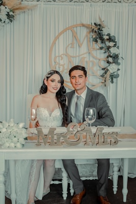 A couple dressed in formal wedding attire sits together at a table. The bride is wearing a white lace dress, and the groom is in a gray suit with a tie. They are surrounded by elegant floral arrangements and a decorative backdrop featuring the phrase 'Mr & Mrs' in wooden letters. Two wine glasses are set on the table in front of them.