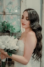 A bride with long wavy hair and elegant makeup holds a bouquet of white roses. She stands near a rustic window with painted floral designs and lush green plants. Her dress is lacey and white, complementing the serene and romantic setting.