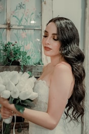 A bride with long wavy hair and elegant makeup holds a bouquet of white roses. She stands near a rustic window with painted floral designs and lush green plants. Her dress is lacey and white, complementing the serene and romantic setting.
