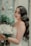 A bride with long wavy hair and elegant makeup holds a bouquet of white roses. She stands near a rustic window with painted floral designs and lush green plants. Her dress is lacey and white, complementing the serene and romantic setting.