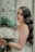 A bride with long wavy hair and elegant makeup holds a bouquet of white roses. She stands near a rustic window with painted floral designs and lush green plants. Her dress is lacey and white, complementing the serene and romantic setting.