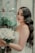 A bride with long wavy hair and elegant makeup holds a bouquet of white roses. She stands near a rustic window with painted floral designs and lush green plants. Her dress is lacey and white, complementing the serene and romantic setting.