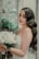 A bride with long wavy hair and elegant makeup holds a bouquet of white roses. She stands near a rustic window with painted floral designs and lush green plants. Her dress is lacey and white, complementing the serene and romantic setting.