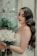 A bride with long wavy hair and elegant makeup holds a bouquet of white roses. She stands near a rustic window with painted floral designs and lush green plants. Her dress is lacey and white, complementing the serene and romantic setting.