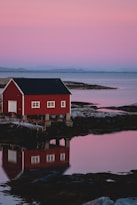 A red wooden cabin stands on stilts by the edge of calm water, reflecting the soft pink hues of a serene sunset in the sky. The peaceful landscape includes distant mountains and rocky shores.
